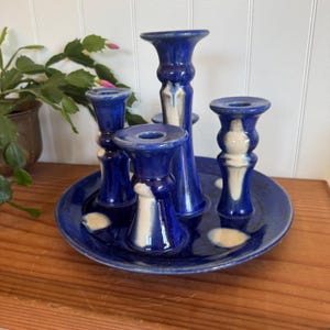May include: A ceramic candle holder set in shades of blue and white. The set includes a round base with four candle holders of varying heights. The candle holders are a deep blue with white accents. The set is on a wooden surface.