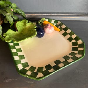 May include: A square ceramic plate with a white center and a green and white checkered border. The plate is decorated with three-dimensional ceramic vegetable accents including a green leaf, a dark blue eggplant, a pink garlic bulb, and an orange carrot.