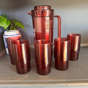 May include: A vintage red cocktail shaker set. The set includes a tall, faceted shaker with a lid and handle, and six matching stackable glasses. The glasses and shaker are a translucent red color.