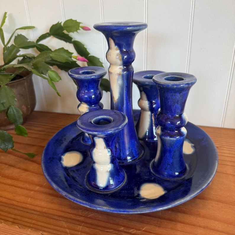 May include: A handcrafted ceramic candle holder with five candle cups. The candle holder is a deep blue with white accents and sits on a matching plate. The design is a unique, decorative piece for any home.