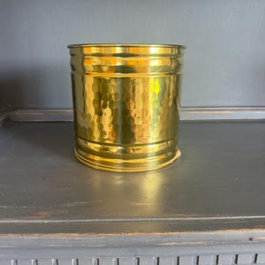 May include: A cylindrical, hammered brass container. The container has a textured surface with a repeating pattern of small, rounded indentations. The brass has a bright, reflective finish, and the container sits on a dark surface.
