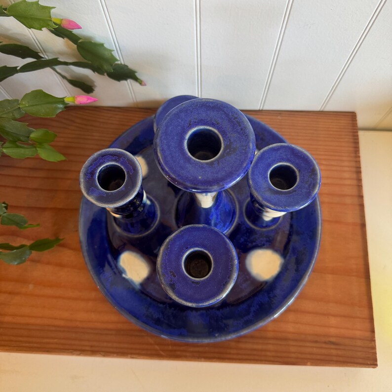 May include: A cobalt blue ceramic candle holder with five circular openings. The candle holder is on a round base with white accents. The piece is placed on a wooden surface.