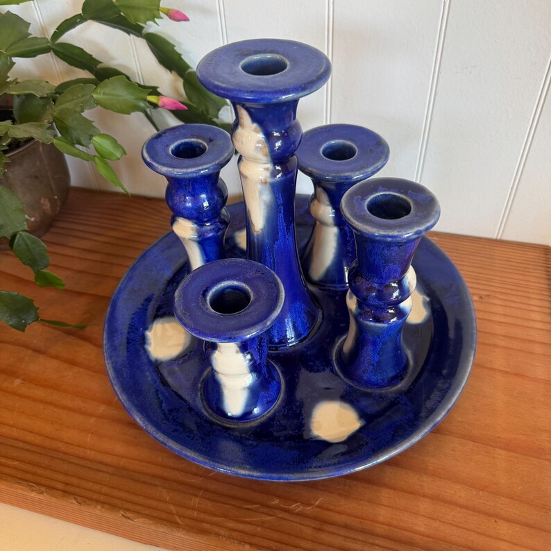 May include: A handcrafted, cobalt blue ceramic candle holder with five candle cups. The candle holder is on a matching blue plate with white accents. The design is a unique, artistic piece for home decor.