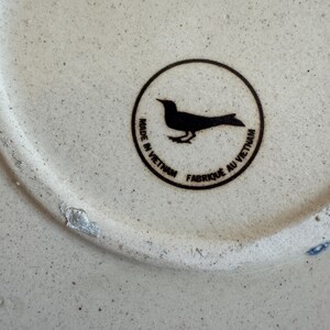 May include: Close-up of a ceramic object with a speckled, off-white surface. A black circular emblem features a bird silhouette and the text "MADE IN VIETNAM" and "FABRIQU&Eacute; AU VIETNAM".