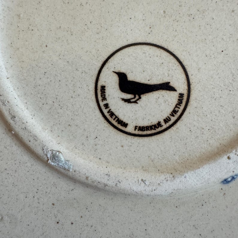 May include: Close-up of a ceramic object with a speckled, off-white surface. A black circular emblem features a bird silhouette and the text "MADE IN VIETNAM" and "FABRIQU&Eacute; AU VIETNAM".