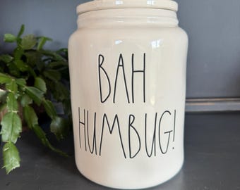 Puede incluir: Un tarro de cerámica de color crema con una tapa a juego que incluye un asa. El tarro tiene una forma cilíndrica con una textura ligeramente irregular y orgánica. El texto BAH HUMBUG! está impreso en letras negras finas y manuscritas.