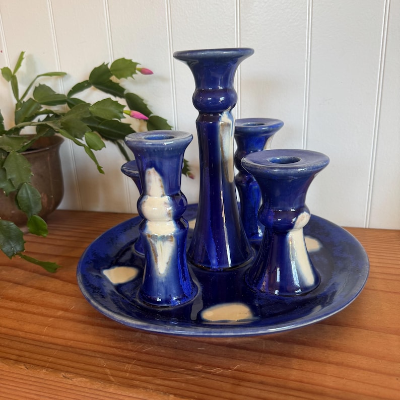 May include: A blue ceramic candle holder with five candle spots. The candle holder is on a round base and has a glossy finish. The candle holder has white accents and is sitting on a wooden surface.