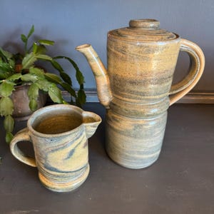 May include: A handmade ceramic coffee pot and creamer set. The coffee pot is tall with a handle and spout, and the creamer has a small handle and spout. Both pieces have a marbled design in shades of tan, gray, and blue. The set is ideal for serving coffee.