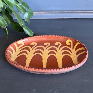 May include: An oval, terracotta-colored ceramic dish with a repeating yellow tree design. The rim has a textured, ridged pattern. The dish is likely used for serving or display.