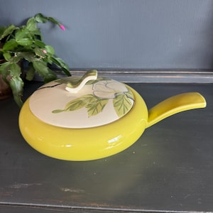 May include: A yellow ceramic serving dish with a lid featuring a floral design. The dish has a handle and a cream-colored lid with a white flower and green leaves. The dish is on a dark surface.