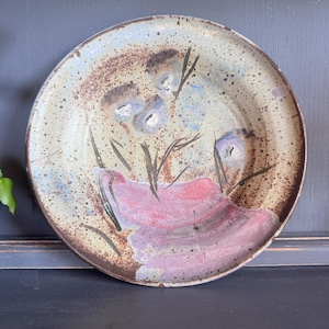 May include: A round ceramic plate with a speckled cream and beige base. The surface features a hand-painted design of abstract purple and blue floral shapes with dark brown stems and a prominent, textured pink wash across the lower section.