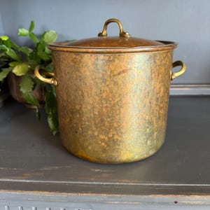 May include: A vintage copper pot with a lid. The pot has a tarnished, mottled finish in shades of gold and green. It features two side handles and a loop handle on the lid. The pot is likely used for cooking or decorative purposes.