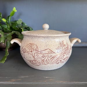 May include: A speckled beige ceramic soup tureen with a lid and handles. The tureen is decorated with a brown illustration of vegetables and a house. The lid has a small round knob.
