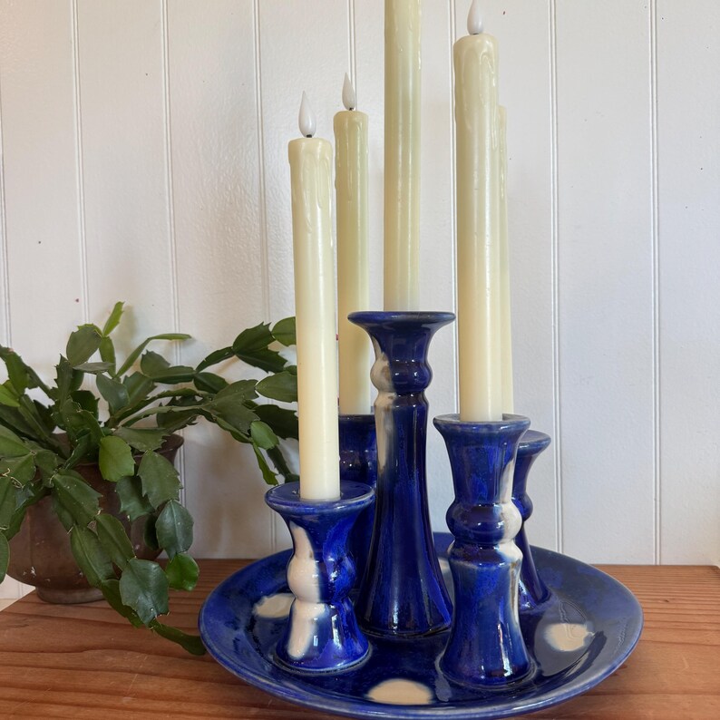 May include: A blue ceramic candle holder with five cream-colored candles. The holder has a circular base with white accents and holds the candles in individual holders. A potted plant is visible in the background.