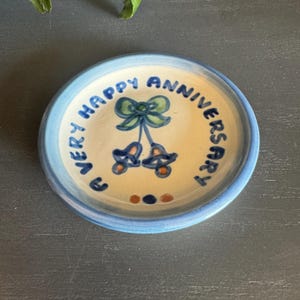 May include: A round ceramic plate with a blue rim and a cream-colored center. The plate features the words "EVERY HAPPY ANNIVERSARY" in blue, along with a green bow and two blue bells with orange accents. A decorative piece.
