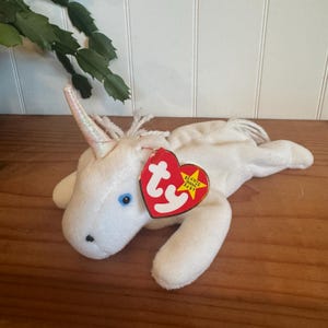 May include: White plush unicorn beanie baby toy with a silver horn and a blue eye. The toy has a red Ty tag with a white star and the words "Beanie Babies Baby".