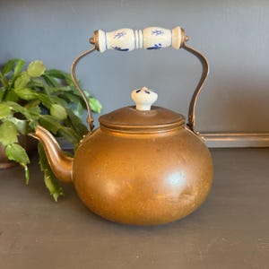 May include: A vintage copper teapot with a white and blue porcelain handle and lid knob. The teapot has a rounded body, a curved spout, and a metal handle. The handle and lid knob feature a blue floral design.