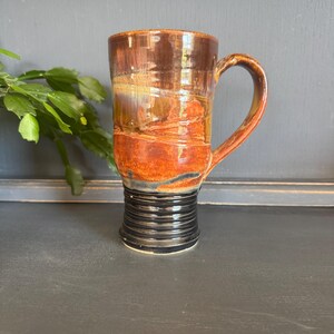 May include: A tall, handmade ceramic mug with a unique design. The mug features a dark brown top that fades into a vibrant orange and black base. The handle is a matching brown color. The base has a textured, ribbed design.