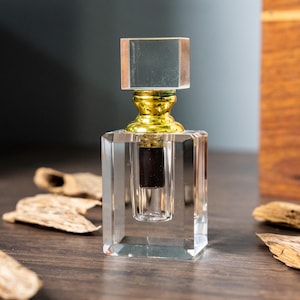 May include: A clear, rectangular perfume bottle with a gold-colored base and a square, clear stopper. The bottle contains a dark brown liquid. The background includes wooden pieces and a wooden box.