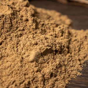 May include: Close-up of a pile of light brown powder with a textured appearance. The powder is on a dark wooden surface, with blurred pieces of bark in the background. The image highlights the texture and color of the powder.