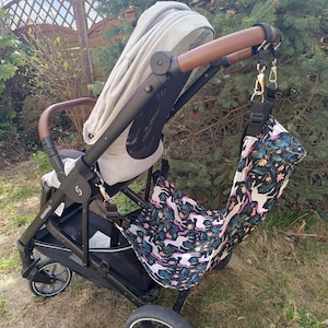 May include: A black and gray stroller with a light gray canopy and brown leather handles. A patterned bag with unicorns hangs from the handle. The bag has a black background with white unicorns and floral accents.