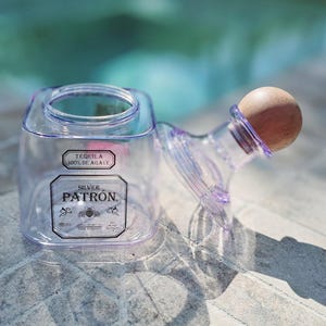 May include: A clear plastic bottle with a wooden stopper. The bottle is labeled "Tequila 100% de Agave" and "Silver Patron".