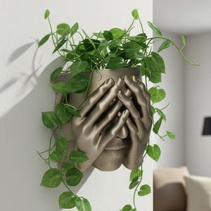 May include: A bronze-colored wall planter shaped like a face with hands covering the eyes. The planter holds green leafy vines that cascade down the wall. The planter is mounted on a white wall, creating a striking contrast.