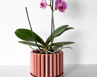 May include: A potted Phalaenopsis orchid with vibrant pink and white petals and dark green leaves, housed in a cylindrical, coral-colored planter featuring a vertical ribbed texture and a matching circular base tray for drainage.