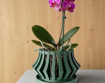 May include: A vibrant magenta orchid with multiple blooms and buds, supported by a green stake, planted in a dark green, slatted, geometric-style plastic pot with a matching circular saucer base, showcasing visible roots and potting medium.