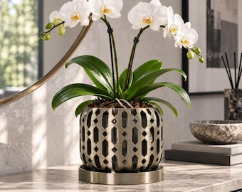 May include: A white orchid plant with green leaves potted in a metallic, cylindrical planter featuring a geometric cutout pattern. The plant is supported by two thin black stakes and sits on a matching circular metal tray base.