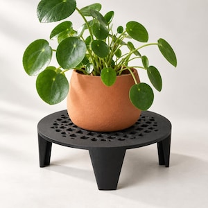 May include: A Pilea peperomioides plant with round green leaves sits in a matte terracotta-colored pot, which is placed on a black circular metal plant stand featuring a geometric grid cutout pattern and three sturdy, angled legs.