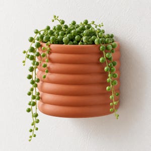 May include: A terracotta-colored wall planter with a ribbed, cylindrical design. The planter is filled with a trailing string of pearls plant, featuring numerous small, round, vibrant green succulent leaves that drape over the sides.