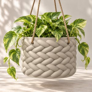 May include: A light beige hanging planter with a raised, braided texture pattern. The cylindrical pot is suspended by three tan rope cords and contains a lush, trailing pothos plant with variegated green and yellow heart-shaped leaves.