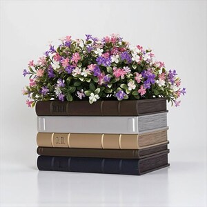May include: A decorative flower pot shaped like a stack of books. The pot is filled with colorful faux flowers in shades of pink, purple, and white. The books are various colors, including brown, gray, and beige.