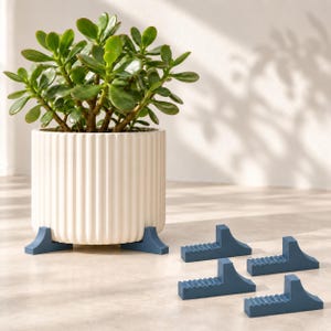 May include: A white, vertically ribbed cylindrical plant pot containing a green jade plant, elevated by three blue, wedge-shaped pot feet. Four additional blue, wedge-shaped pot feet with a ridged top surface are arranged nearby.