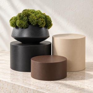 May include: A matte black bowl-shaped planter containing a cluster of green succulent-like plants, resting on a black cylindrical pedestal. Beside it are two additional cylindrical pedestals, one in a dark brown finish and one in a light beige finish.