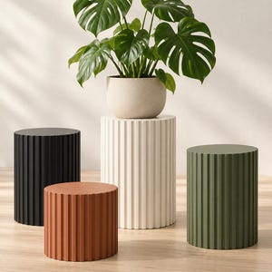 May include: A collection of four cylindrical, fluted pedestals in black, terracotta, white, and olive green. The tallest white pedestal supports a potted Monstera plant with large, split green leaves in a speckled, light-colored round planter.