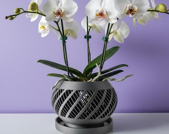 May include: A white orchid plant with multiple blooms and green buds, supported by thin green stakes, potted in a dark gray, spherical planter featuring a diagonal lattice pattern and a matching circular drip tray at the base.