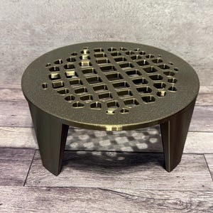 May include: A bronze-colored, round, decorative drain cover with a geometric pattern. The drain cover is supported by three angled legs. The design features a series of rectangular and hexagonal cutouts, creating an open, airy appearance.