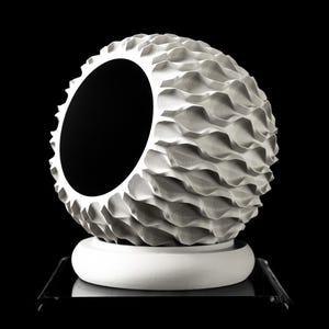 May include: White ceramic sculpture with a textured, wavy surface. The sculpture is shaped like a sphere with a hole in the center. It is displayed on a white pedestal.
