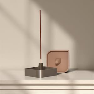 May include: A modern incense holder set. The set includes a square, metallic gray incense holder, a light brown incense stick, and a matching light brown, square-shaped incense ash catcher.