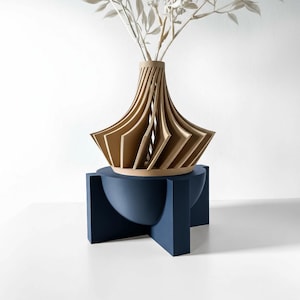 May include: A modern, geometric vase with a layered, fan-like design in shades of tan and beige. The vase sits atop a navy blue, pedestal-style stand. The vase holds white, dried floral stems.