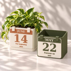 May include: Two square planters designed like calendar pages. The left planter is cream with a terracotta accent, reading JUNE 14, Our beginning, 2024, holding a pothos plant. The right planter is olive green, reading MAY 22, Forever us, 2025.