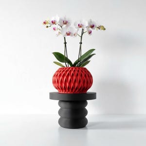 May include: A red geometric planter holding white orchids with purple centers, set on a black, stacked-cylinder plant stand. The modern design features a striking contrast of colors and textures, suitable for home decor.