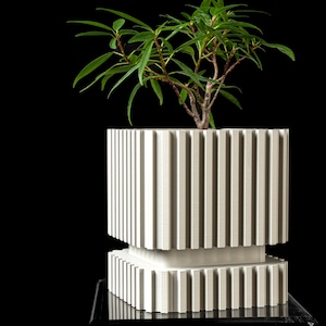 May include: A white square planter with a vertical ribbed texture featuring a recessed middle section. The planter has a two-tiered design with a wider top section and a narrower base. A small green leafy plant grows out of the top of the planter.