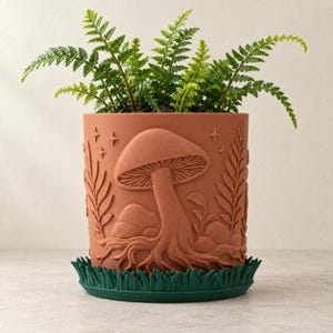 May include: A terracotta-colored cylindrical plant pot featuring a raised relief design of a large mushroom, ferns, and small stars. The pot sits inside a dark green saucer molded to resemble blades of grass, containing a green fern plant.
