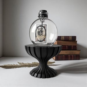 May include: A clear, round glass bottle with a black stopper and a decorative label, displayed on a black pedestal. The label features an illustration and the text "RAT SALIVA". The pedestal has a unique, fluted design.