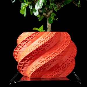 May include: A bright orange planter with a spiraling, geometric, textured surface pattern. The cylindrical container features a repeating triangular lattice design across its exterior. A small green plant with dark leaves grows from the top.