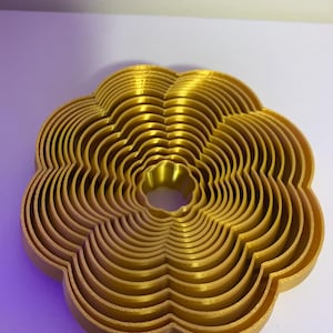 Puede incluir: Objeto dorado en forma de flor, compuesto por múltiples capas. El diseño presenta anillos concéntricos y festoneados, creando un efecto 3D. Tiene un agujero central y una superficie texturizada. Probablemente un objeto decorativo.