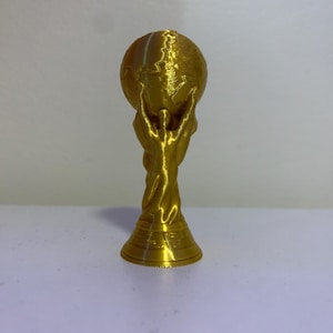 Puede incluir: Un trofeo dorado que representa un globo terráqueo sostenido por dos figuras estilizadas. El trofeo tiene un diseño detallado y una base circular. La forma general es simétrica y el acabado es brillante.